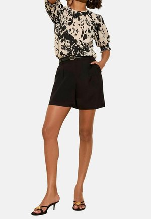 Femme portant un chemisier noir et beige à imprimé animalier avec manches bouffantes, un short noir ceinturé et des sandales à talons noires avec un détail doré.