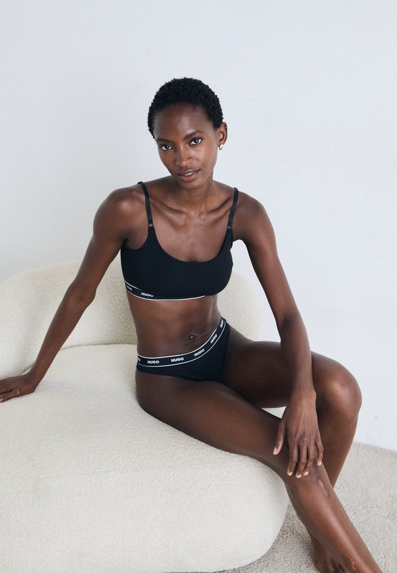 Woman with short curly hair sitting on a white curved chair wearing black Hugo branded underwear and a matching bra, against a plain background.