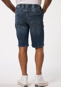 Pantalones cortos de mezclilla en azul oscuro con una cinturilla elástica. Incluye dos bolsillos traseros y un dobladillo enrollado, rematados con zapatillas blancas.