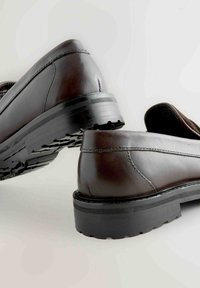 Brązowe skórzane buty z okrągłym czubkiem, niskim obcasem i teksturowaną podeszwą. Szwy widoczne wzdłuż boków, ukazujące gładkie wykończenie.