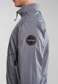 GrauJacke mit hohem Kragen, ausgestattet mit einem runden Logo-Patch auf dem linken Ärmel, seitlichen Reißverschlusstaschen und einer glatten, glänzenden Textur.