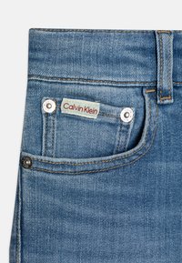 Primer plano de un bolsillo de jeans de denim azul con etiqueta beis de Calvin Klein y remaches de metal sobre un fondo claro.