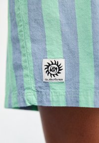 Gestreifte Stoffshorts in hellgrün und blau, mit einem gestickten Logo-Patch mit einem Sonnendesign. Das Material wirkt leicht und lässig.