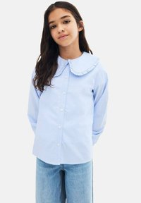 Vaaleansininen, raidallinen napitettava paita, jossa on suuri röyhelökaulus ja pitkät hihat. Valmistettu sileästä kankaasta. Rentomallinen, yhdistetty farkkuihin.