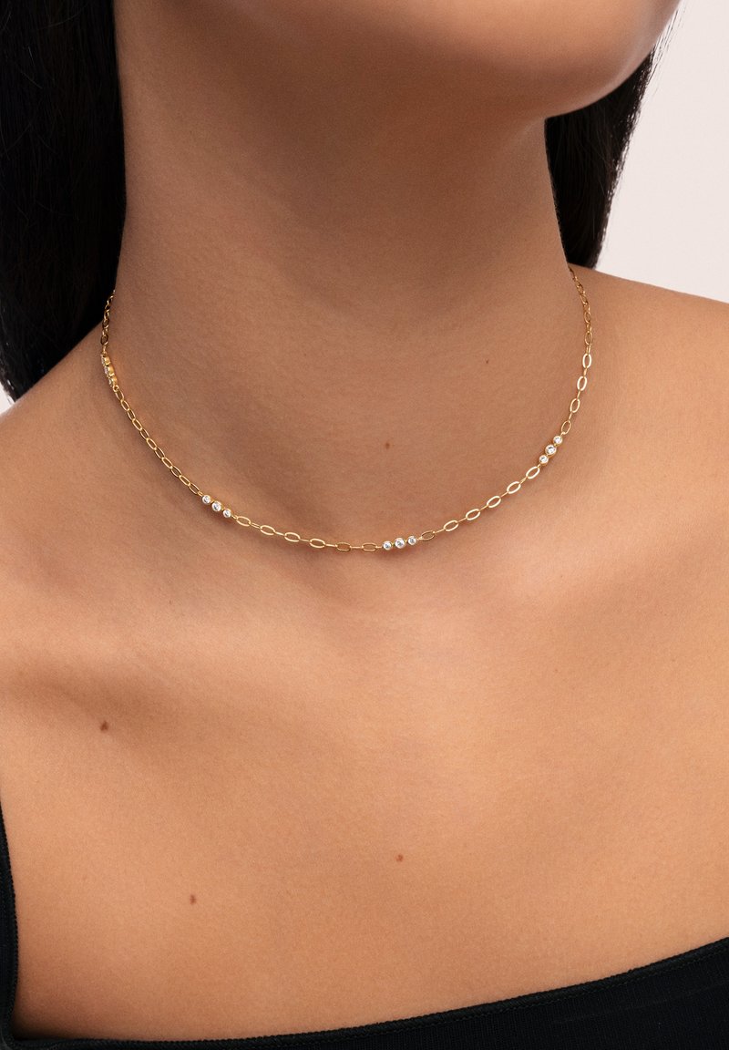 Collar de cadena dorada con eslabones ovalados, adornado con tres pequeños acentos brillantes. Se lleva ajustado al cuello sobre piel desnuda.
