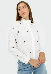 Giovane donna che indossa una camicia bianca con bottoni decorata da piccoli motivi floreali rossi e jeans azzurri, che guarda di lato.