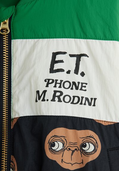 Πολύχρωμο μπουφάν με πράσινες, λευκές και μαύρες πινελιές. Ο μπροστινός σχεδιασμός περιλαμβάνει το κείμενο "E.T. PHONE M. RODINI" και μια εικονογραφημένη φιγούρα. Χρυσός φερμουάρ.
