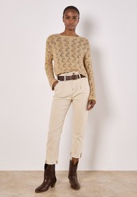 Beigefarbener Strickpullover mit Spitzenmuster, cremfarbene Hose, brauner Gürtel und braune Knöchelboots. Fokus auf Textur und Details in der Kleidung.