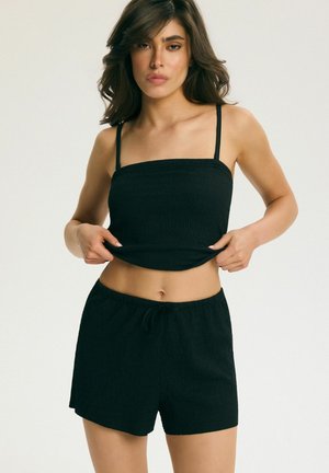 Kobieta o włosach do ramion, ubrana w czarny, bezrękawnikowy crop top i pasujące czarne szorty, poprawiająca dół topu.