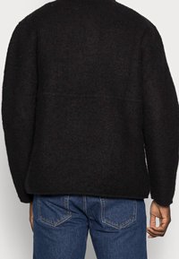 Mustavillainen villapaita, jossa on rento istuvuus ja laskeutuvat olkapäät, suora helma ja minimaalinen saumaus. Yhdistetty sinisiin farkkuihin.