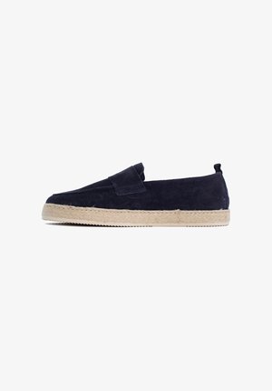 Slip-on schoen in marineblauw suède met een beige gevlochten espadrille-achtige zool en een lusje aan de hiel, getoond in profiel op een witte achtergrond.