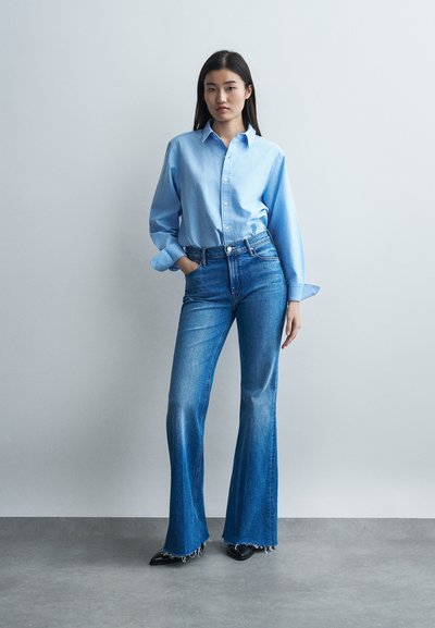 Žena stojaca pri hladkej stene, oblečená v svetlomodrej košeli na zapínanie na gombíky a vo vysokým pásom rozšírených džínsach s rozstrapkanými lemami a čiernymi špicatými topánkami.