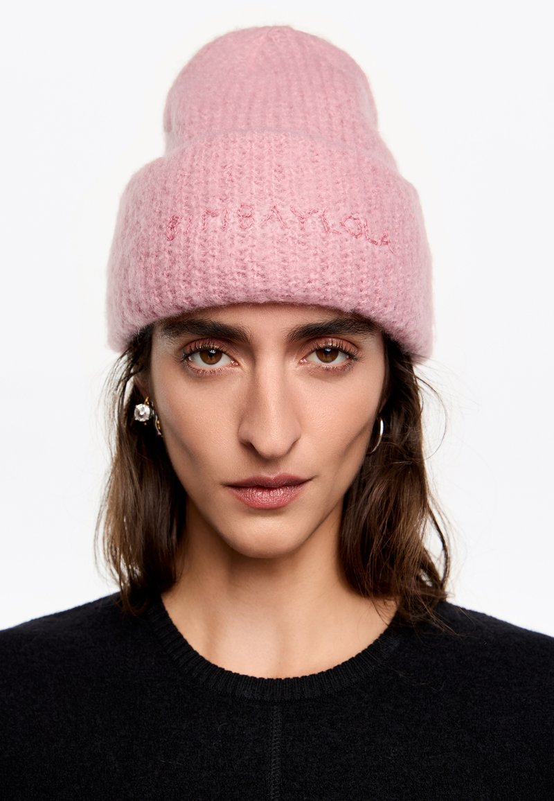 Różowy dzianinowy beanie o miękkiej fakturze, z zaokrąglonym szczytem i haftowanym tekstem. Noszony z czarnym swetrem, minimalistyczny design.