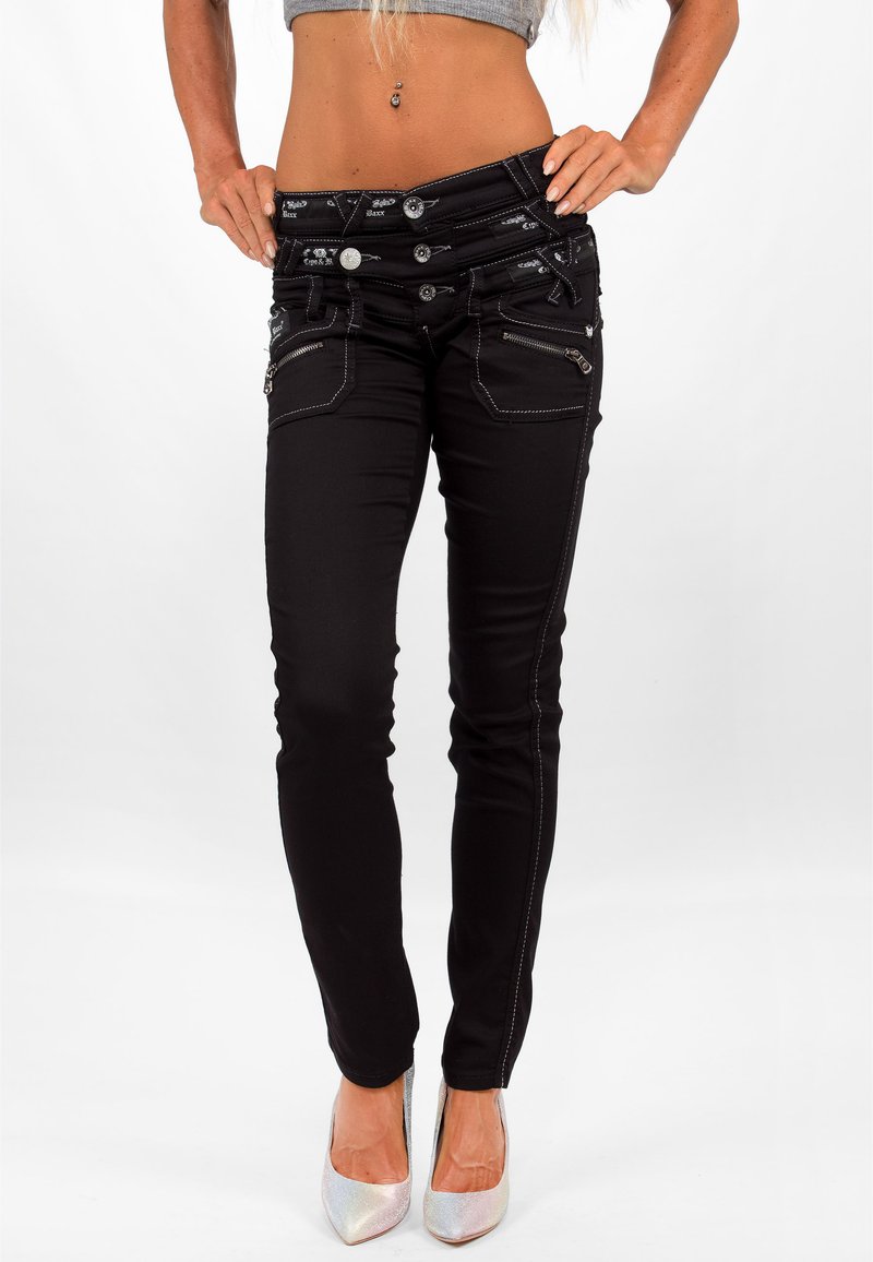 Zwarte skinny jeans met een hoge taille, zilveren stiksels, meerdere ritszakken en decoratieve knoopaccenten. Gekleed met zilveren hoge hakken.