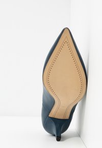 Chaussure à talon haut en cuir bleu avec un bout pointu et un design minimaliste. Texture lisse et semelle marron clair avec un détail en motif triangulaire.