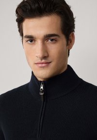 Ung man med mörkt hår som bär en svart ribbad zip-up-tröja med hög krage, neutral bakgrund.