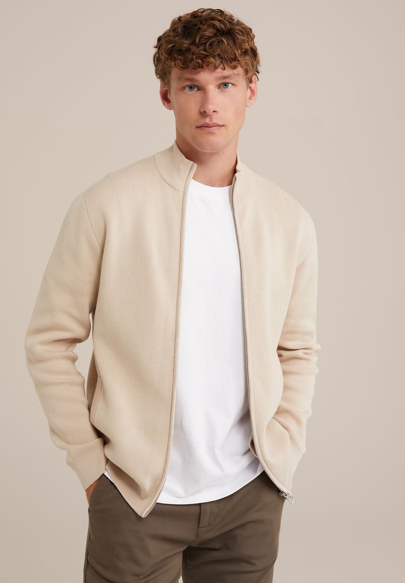 Bēša rāvējslēdzēja kardigāns, izgatavots no adīta auduma, ar stāvošu apkakli, ribinātām piedurknēm un gludu tekstūru, uzvilkts virs baltas T-kreklu.