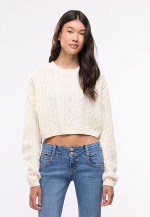 Giovane donna con lunghi capelli scuri che indossa un maglione corto bianco a trecce e jeans blu a vita alta su uno sfondo semplice.