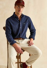 Námornícka košeľa s gombíkmi a náprsným vreckom, s výšivkou loga, skombinovaná s smotanovými manžestrovými nohavicami a hnedými koženými topánkami. Marhuľová čiapka na hlave.