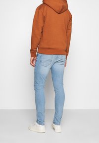 Muž v světle modrých úzkých džínách, bílých teniskách a rezavě zbarvené mikině s kapucí stojí zády k divákovi na jednobarevném pozadí.