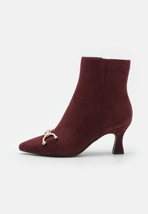 Bottine en daim rouge foncé avec bout pointu, talon bas incurvé et boucle décorative dorée près de l'avant sur un fond uni.