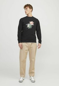 Sudadera de algodón negra con diseño gráfico floral que presenta elementos en rosa y verde, combinada con pantalones beiges y zapatillas plateadas.