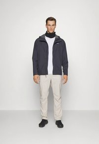 Αδιάβροχο σκούρο γκρι μπουφάν με κουκούλα, λευκό πουκάμισο, ανοιχτό μπεζ παντελόνι και μαύρα παπούτσια. Το μπουφάν διαθέτει μπροστινό φερμουάρ και ρυθμιζόμενα κορδόνια.