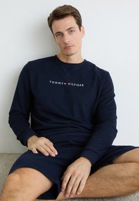 Mies, jonka lyhyet ruskeat hiukset ja siniset silmät, pukeutuneena tummansiniseen Tommy Hilfiger -sweatshirt-paitaan ja samansävyisiin shortsseihin, istuu valkoista seinää vasten.