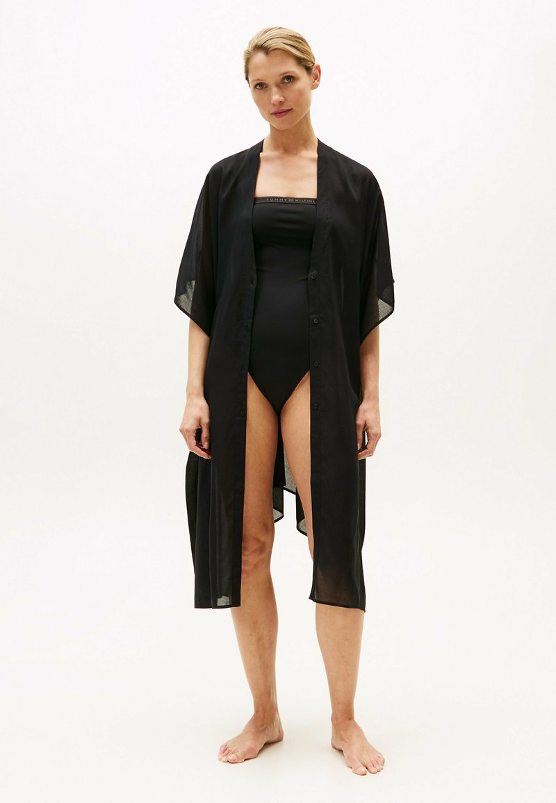 Copricostume nero in stile kimono trasparente con maniche corte, apertura frontale e chiusure con bottoni. Indossato sopra un costume da bagno nero, presenta una texture leggera.