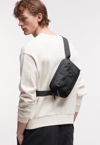 Borsa a tracolla in nylon nero con doppi zip, indossata sopra una felpa beige chiaro, dotata di una tracolla piatta e una forma strutturata.