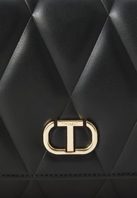 Czarna torebka z sztucznej skóry z pikowanym diamentowym wzorem, z metalowym zapięciem w kolorze złotym i wygrawerowanym napisem "TWINSET". Kompaktowy i zorganizowany design.
