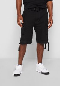 Mężczyzna ubrany w czarne szorty cargo z bocznymi kieszeniami i białe sneakersy Nike wysokie, stojący na jednolitym szarym tle.