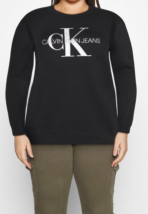 Osoba ubrana w czarną bluzę Calvin Klein Jeans z dużym białym logo CK oraz oliwkowozielone spodnie na jednolitym tle.