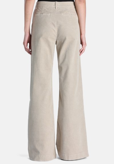 Personne portant un pantalon large en velours côtelé beige taille haute, vue de dos sur fond blanc uni.
