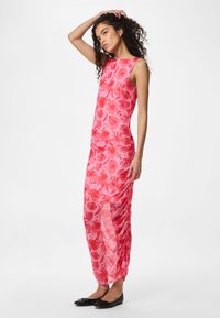 Ženska s kodrastimi lasmi, oblečena v rdečo cvetlično maxi obleko brez rokavov in črne balerinke, stoji z eno roko na glavi.