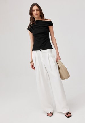Kobieta ubrana w czarny top z odkrytymi ramionami, szerokie białe spodnie, czarne sandały, trzyma beżową torebkę na jednolitym jasnym tle.