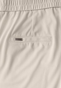 Tissu beige clair avec une texture lisse, comportant une taille élastique et une poche cousue avec une étiquette métallique.