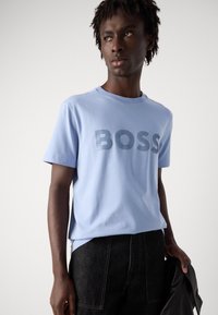 Šviesiai mėlynas medvilninis marškinėliai su ryškiai mėlynu "BOSS" logotipu, apvaliu iškirpimu ir trumpomis rankovėmis, derinami su tamsiai mėlynomis džinsinėmis kelnėmis.