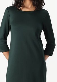 Nainen, joka käyttää tummanvihreää pitkähihasta crew neck -mekkoa, kädet selän takana valkoista taustaa vasten.