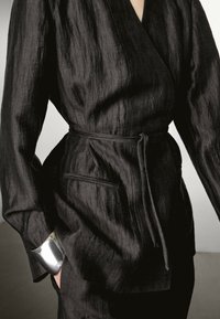 Schwarze strukturierte Wickeljacke mit einem taillierten Gürtel, langen Ärmeln und einer Seitentasche. Verfügt über einen glatten, schimmernden silbernen Akzent an den Bündchen.