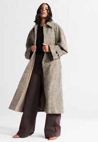 Long manteau en tweed marron à motif à carreaux, avec un large col, coupe décontractée et fermeture par boutons, superposé sur un pantalon marron foncé.