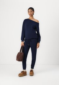 Felpa blu navy off-shoulder con maniche lunghe, abbinata a pantaloni della tuta coordinati e sneakers con piattaforma marroni. Borsa a tote marrone in mano.