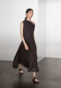 Donna che cammina all'interno, indossando un top scuro monospalla, una gonna midi fluente, sandali neri senza tacco e orecchini pendenti.