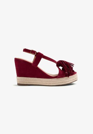 Sandalia de cuña de ante rojo con hebilla en la correa del tobillo, suela plataforma de yute trenzado y detalle de borla en la correa delantera.