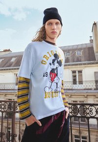 Svijetloplava majica s kratkim rukavima s grafikom Mickey Mouse, složena preko prugaste majice s dugim rukavima. Tamne hlače i crna kapa za glavu upotpunjuju izgled.