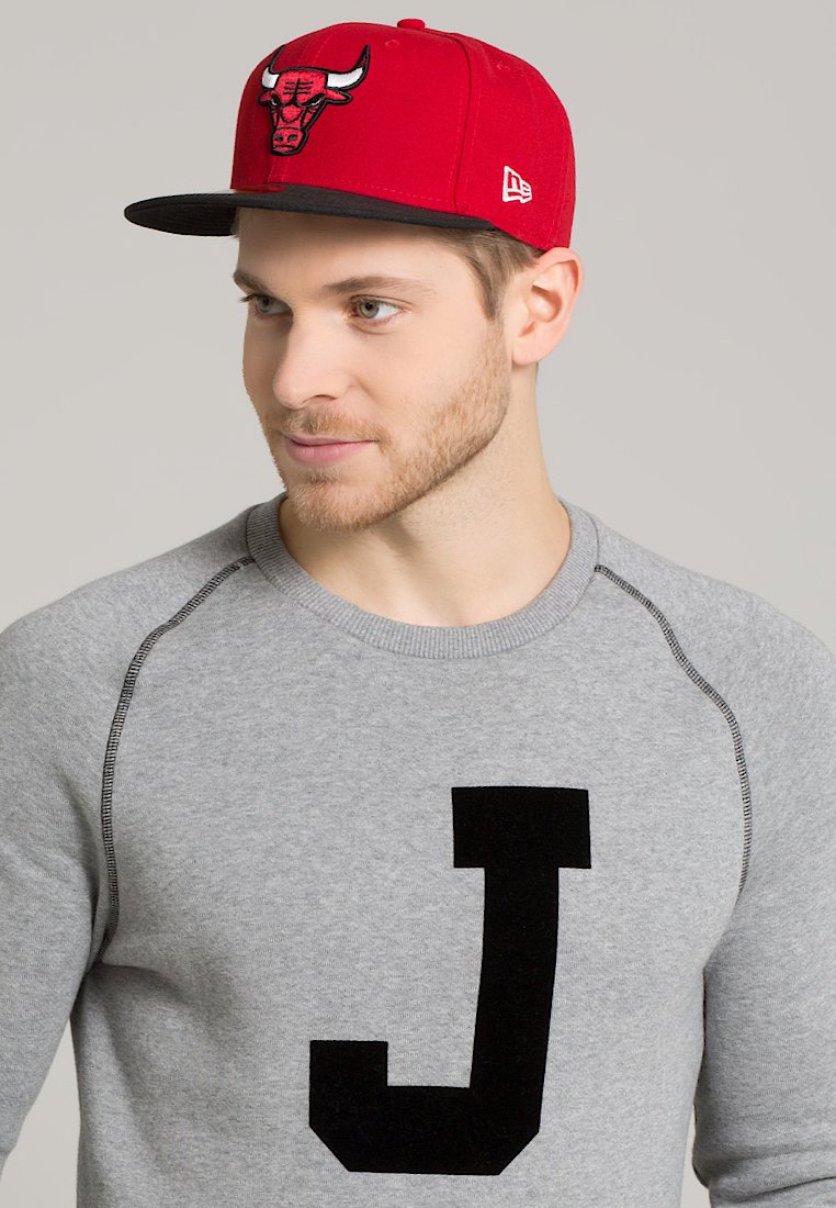 Gorra de béisbol roja con visera negra que presenta un logo de toro bordado; sudadera gris con una gran letra "J" negra en el frente.