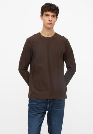 Homme aux cheveux bouclés portant une chemise marron à manches longues et un jean bleu, debout devant un fond blanc uni avec les mains derrière le dos.