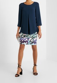 Blouse marine à manches trois-quarts avec détail de plis, associée à une jupe à motifs aux couleurs pastel et à une écriture, complétée par des escarpins à talons carrés noirs.