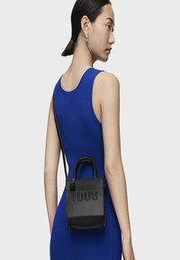 Bolso tote de cuero negro con el logo "TOUS" en relieve, asa corta y correa ajustable, que presenta un pequeño detalle decorativo de pompon.