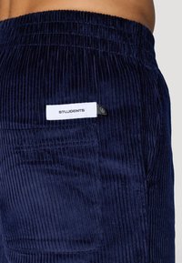 Mørkeblå kordfløyelsbukser med vertikale ribber, med en hvit etikett på baklommen. Elastisk midje, glatt tekstur og uformelt design.